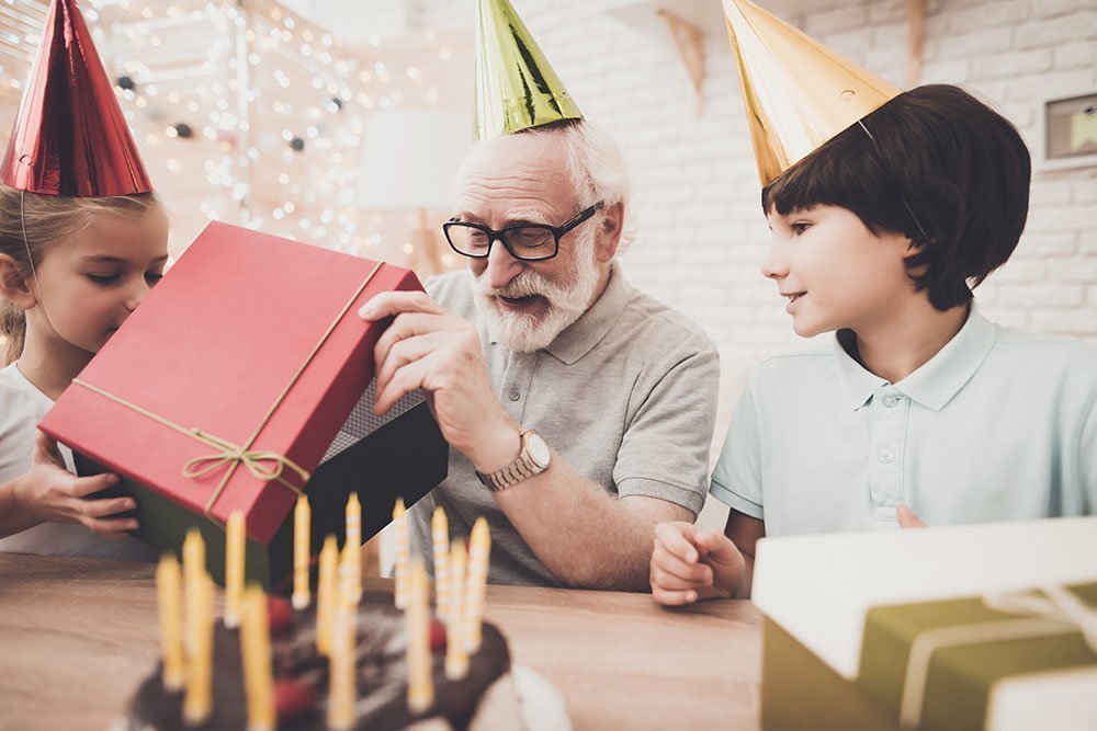 El abuelo recibe un regalo de cumpleaños de dos nietos detrás de un pastel con velas.